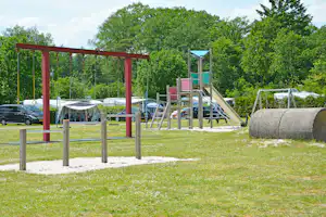 Spielplatz mit Schaukeln und Rutsche auf Sandfläche, im Hintergrund Zelte und geparkte Autos, umgeben von Bäumen.