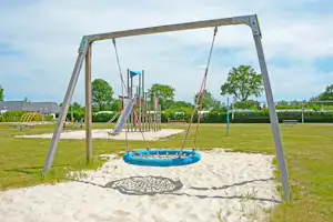 Schaukel auf dem Sandspielplatz mit Seilaufhängung, Rutsche und Spielgeräten im Campingpark, im Hintergrund Bäume und Gebäude.