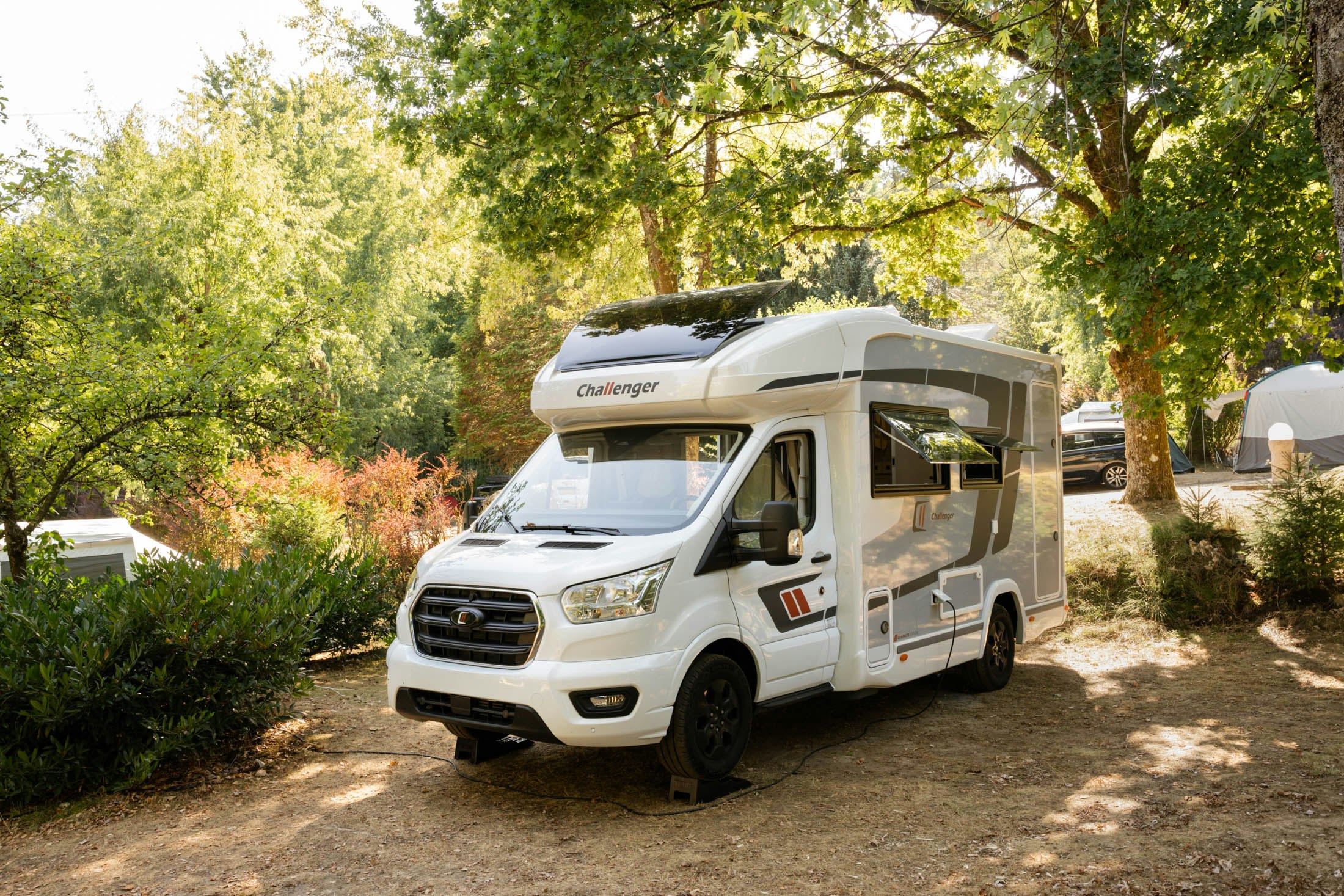 Wohnmobil auf einem Campingplatzweg zwischen Bäumen, mit geöffneter Markise und weißem Aufbau.