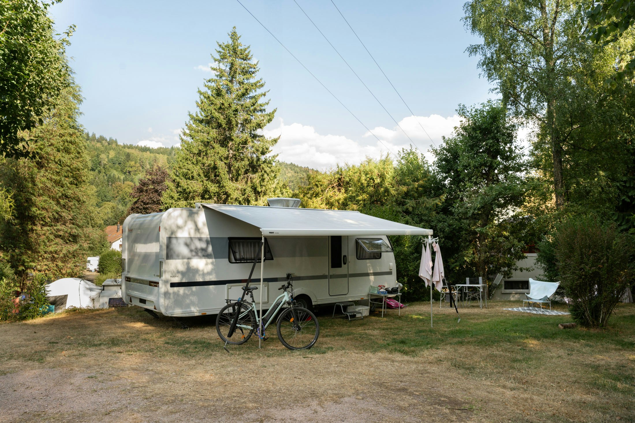 Wohnwagen mit Vorzelt und Fahrrad am Stellplatz, umgeben von Bäumen und Wiesen, im Hintergrund Hügel und Berge.