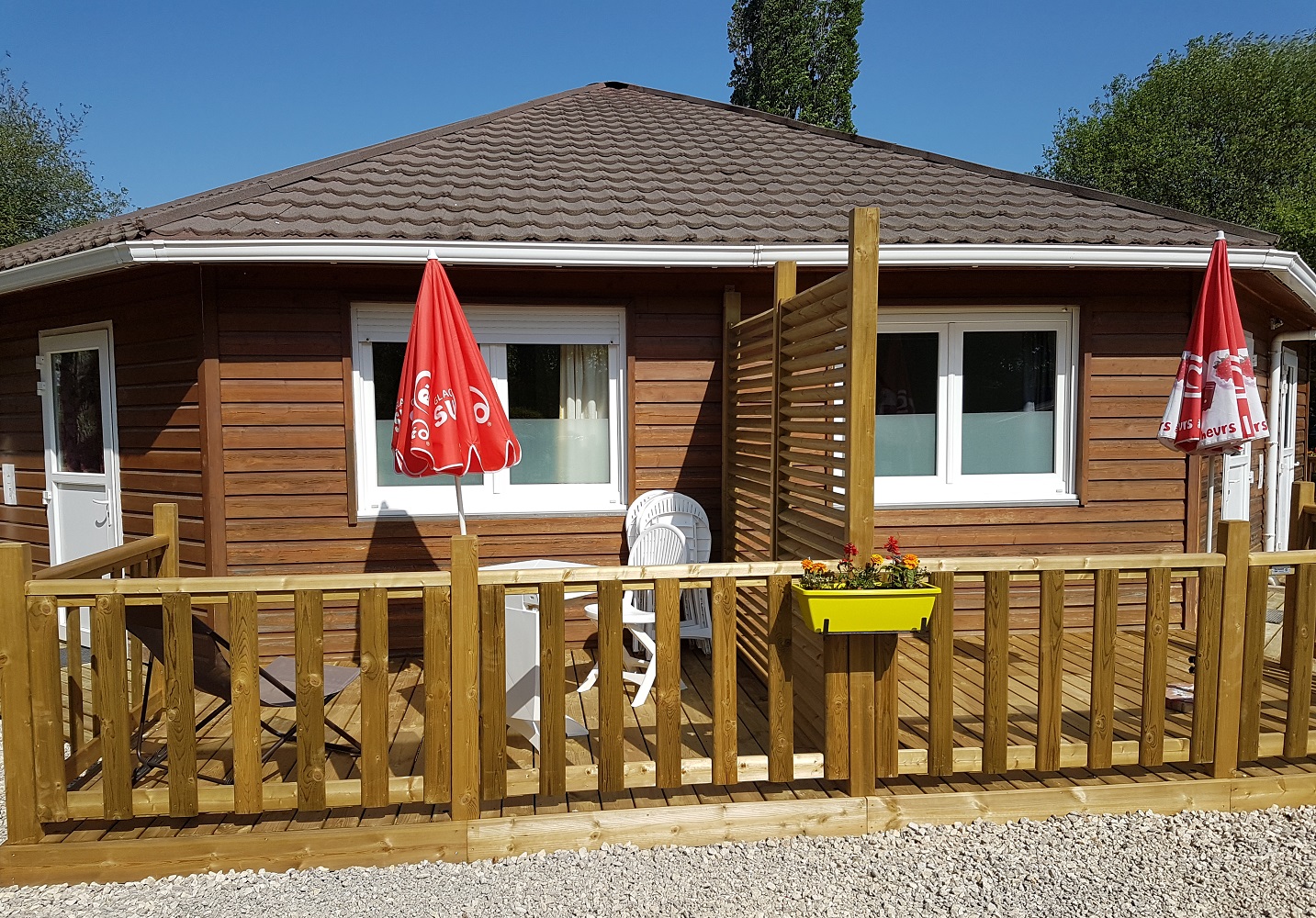 Holzbungalow mit Terrasse und Holzgeländer, zwei rote Fahnen am Eingang, Fenster und Sitzbank vor dem Gebäude.