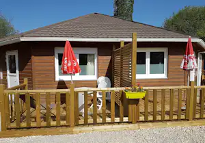Holzbungalow mit Terrasse und Holzgeländer, zwei rote Fahnen am Eingang, Fenster und Sitzbank vor dem Gebäude.