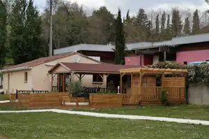 Bungalow mit Terrasse und Holzsichtschutz, davor Rasenfläche; im Hintergrund Bäume und ein weiteres Gebäude auf dem Campingplatz.