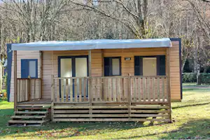 Holz-Blockhaus mit Terrasse und Geländer im Campinggelände, umgeben von Bäumen und Rasen.