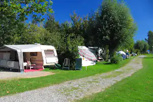 Wohnwagen mit Vorzelt auf einem Campingplatz, daneben Stühle und Tisch, Reihe weiterer Fahrzeuge und Zelte im Grünen.