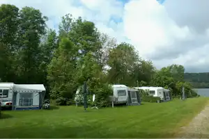 Campingplatz mit Wohnwagen und Zelt auf grünem Rasen, umgeben von Bäumen und einem Weg.