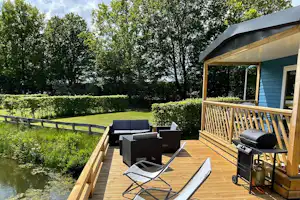 Camping De Papillon - Blick auf Standplätze und moderne Mietunterkunft mit überdachter und Sonnenterrasse, Sitzgelegenheit und Grill direkt am Teich gelegen