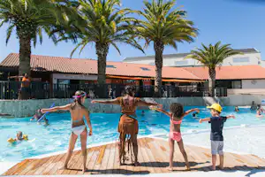 Kinder stehen am Pool im Campingbereich, Holzdeck, Palmen und mehrere Personen im Wasser vor Gebäuden mit roten Dächern.