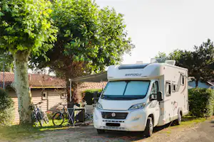 Weißes Wohnmobil am Campingplatz mit Fahrrädern am Zaun, Bäumen und Häusern im Hintergrund.