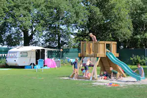 Wohnmobil am Campingplatz mit Vorzelt, daneben Spielplatz mit Holzspielgerüst und grünem Rutschbahn auf Rasen.