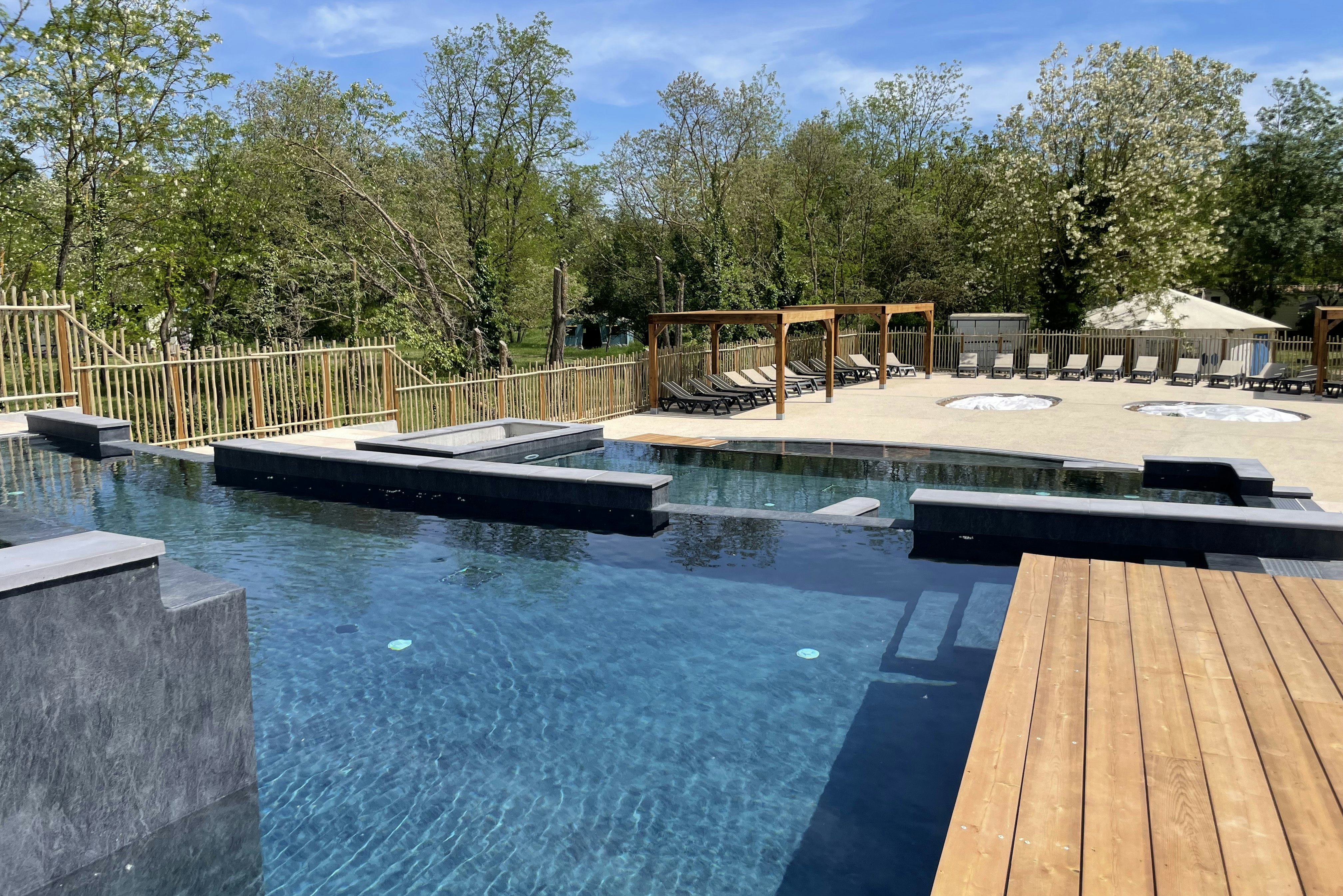 Camping-Pool mit Liegen auf der Terrasse, Holzgeländer und Zelte im Hintergrund, umgeben von Bäumen und Grün.