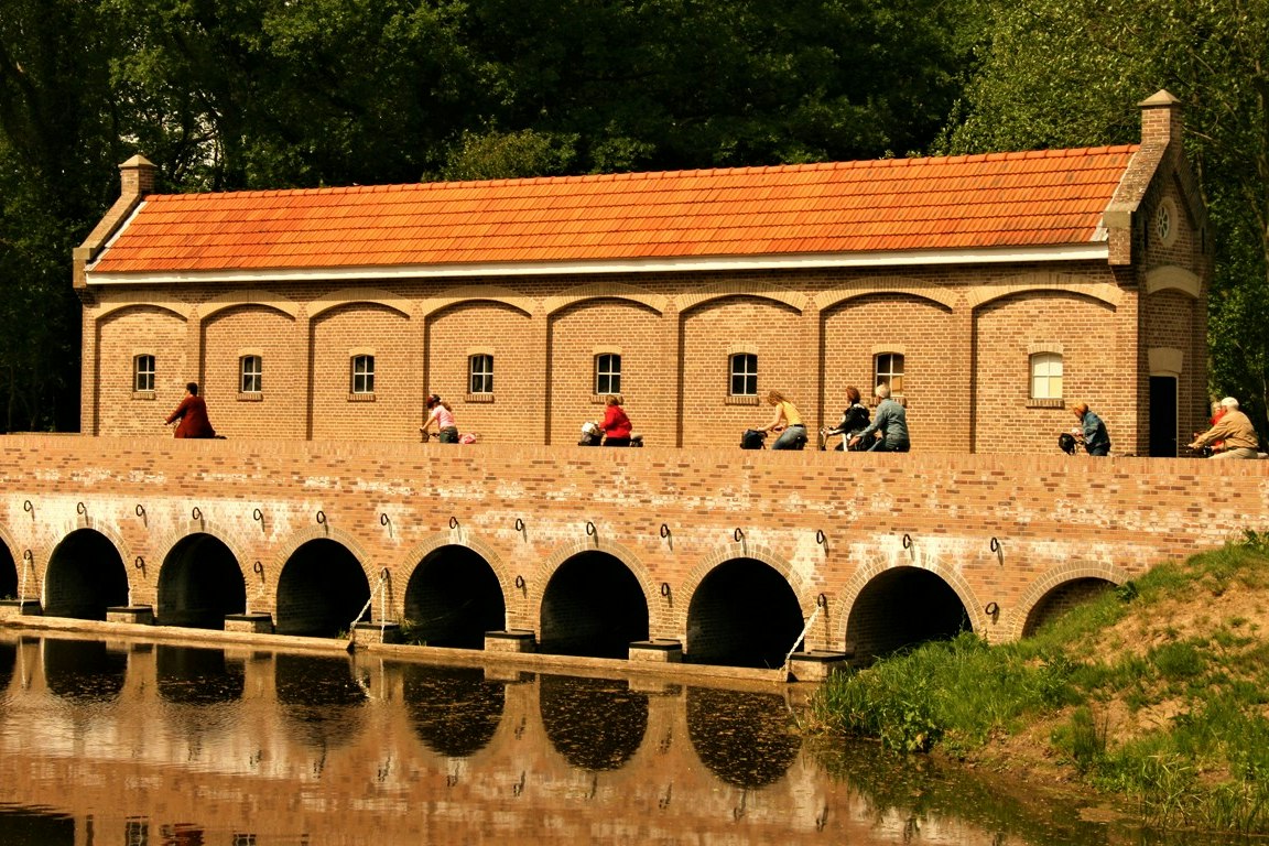 Backsteingebäude mit orangefarbenem Dach am Kanal, Bögen und Fensterfront, davor Wasser und Gras am Ufer.