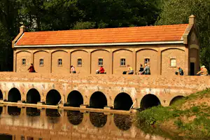 Backsteingebäude mit orangefarbenem Dach am Kanal, Bögen und Fensterfront, davor Wasser und Gras am Ufer.