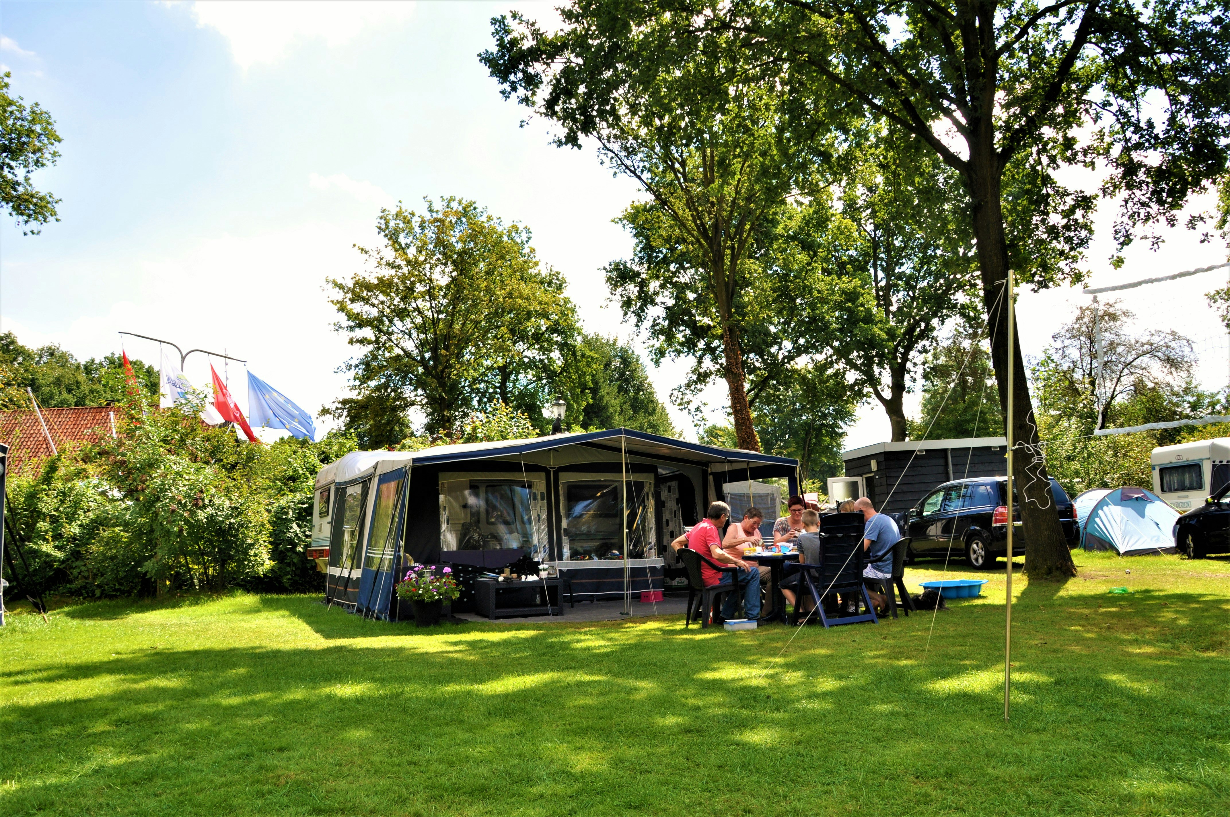 Wohnwagen mit Vorzelt und Terrasse auf dem Campingplatz, daneben Autos, Zelte und Bäume im grünen Garten.