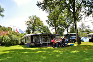 Wohnwagen mit Vorzelt und Terrasse auf dem Campingplatz, daneben Autos, Zelte und Bäume im grünen Garten.