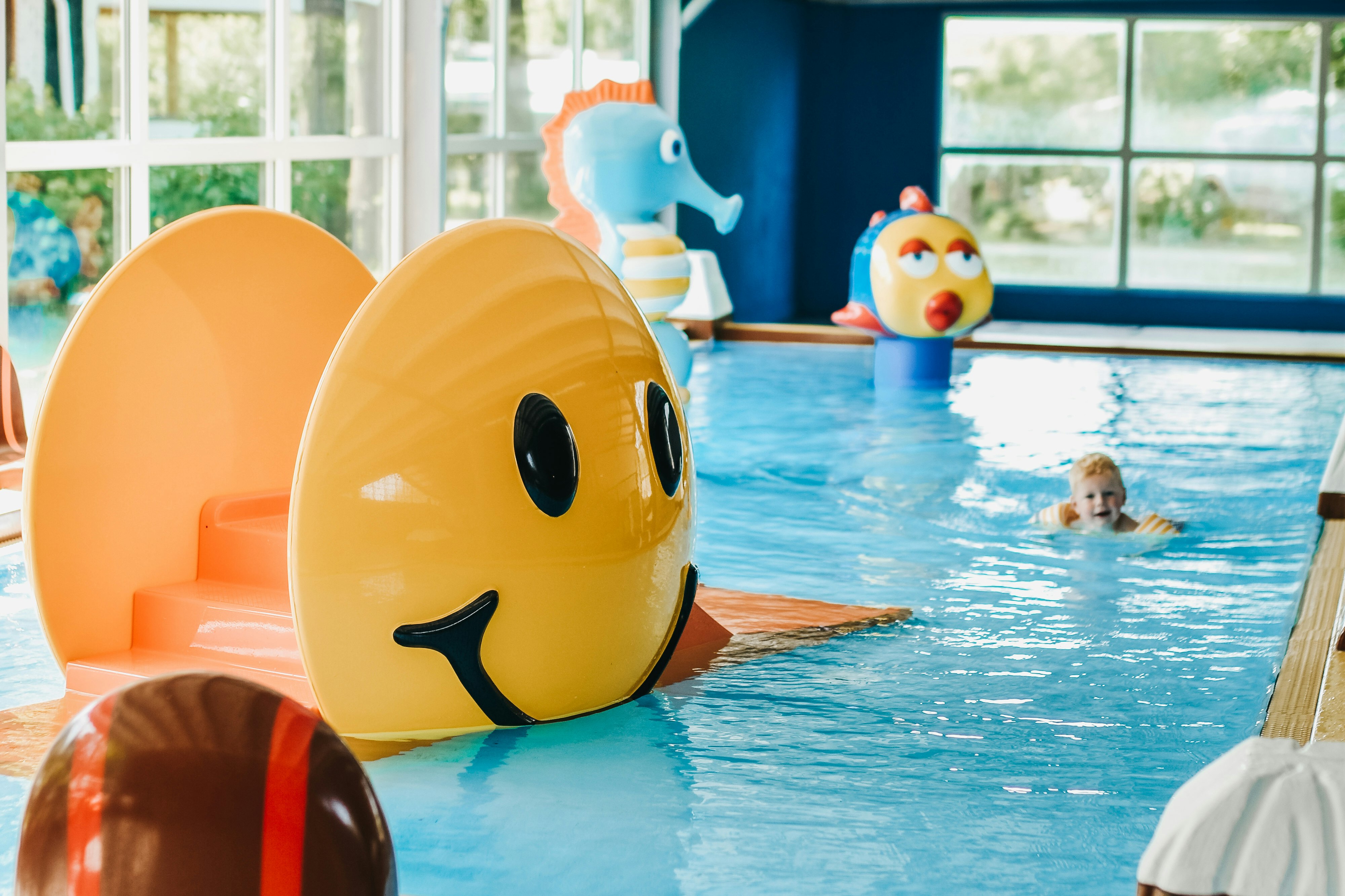 Gelbe und orange Schwimmtiere auf dem Beckenrand, im Hintergrund ein Kind im Kinderbecken und große Fensterfront.