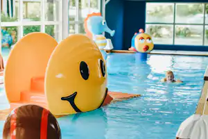 Gelbe und orange Schwimmtiere auf dem Beckenrand, im Hintergrund ein Kind im Kinderbecken und große Fensterfront.