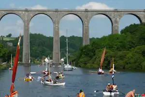 Segelboote auf dem Fluss unter einer Steinbogenbrücke, im Hintergrund Bäume und weitere Boote im Wasser.