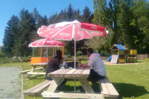 Camping-Tisch mit zwei Personen unter roten Sonnenschirmen, im Hintergrund Spielplatz und Bäume im Grünen.