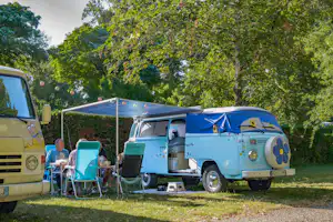 Blauer VW-Bus mit Markise und Vorzelt, daneben Campingstuhl und Tisch auf dem Stellplatz im Grünen.