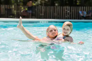 Zwei Kinder und ein Erwachsener im Schwimmbecken, im Hintergrund Zaun und Bäume im Campingbereich.