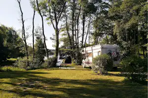Weißes Wohnmobil auf einem Campingplatz zwischen hohen Bäumen, mit Wiese und Blick auf einen Fluss im Hintergrund.