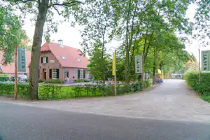 Weg im Campinggelände zwischen Bäumen, dahinter ein Gebäude mit rotem Ziegeldach und Fensterfronten.
