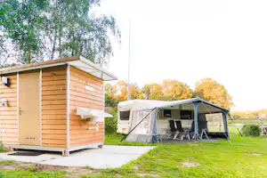 Wohnmobil mit Vorzelt und Campingstühlen neben einer Holzblockhütte mit Tür, Waschbecken und Wasserhahn auf dem Stellplatz.