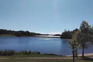 Blick auf einen See mit Badebojen, Uferweg und Bäumen am Rand, im Hintergrund bewaldete Hügel.