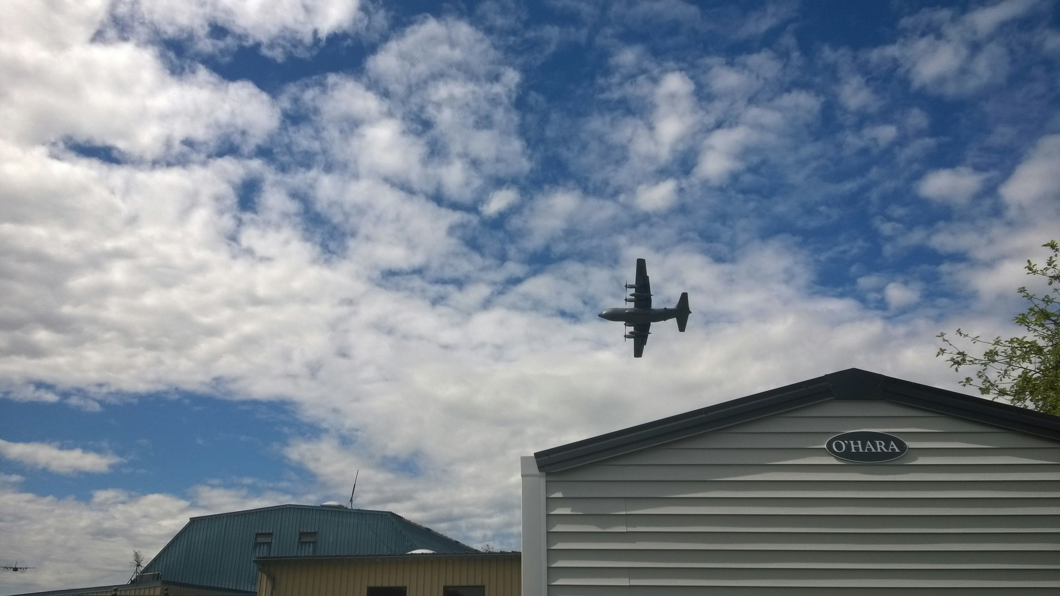 Flugzeug am Himmel über einem Campingplatz mit Gebäude und Schriftzug „O’HARA“ unter bewölktem Himmel.