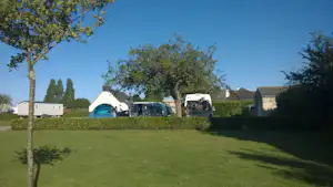 Campingwiese mit Wohnwagen und Zelt im Hintergrund, umgeben von Bäumen und Hecken, im Blick Richtung Gebäude.