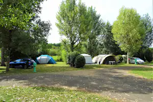 Zelte auf einem Campingplatz zwischen Bäumen, mit Autos am Stellplatz und Heckenbeeten im grünen Gras.