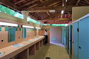 Helles Sanitärgebäude mit Waschbecken an einer langen Theke, Fliesenboden und offenen Türen, im Hintergrund eine Person.