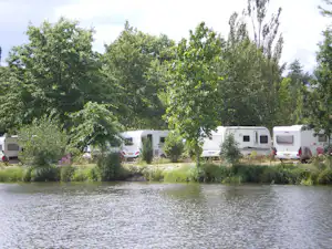 Wohnmobile und Campingbusse stehen am Flussufer im Campingplatz, umgeben von Bäumen und grünem Uferbewuchs.