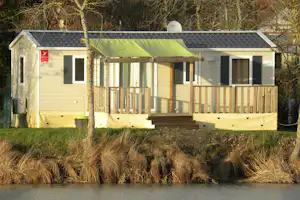 Mobilheim mit Terrasse am Flussufer, umgeben von Bäumen und Gras, im Campingplatzbereich.