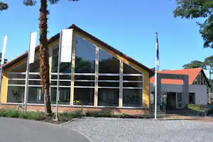 Campinggebäude mit großer Glasfront, Eingang und Fahnen vor dem Haus, gepflasterter Weg und Kiesfläche.