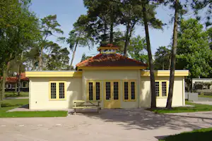 Weißes Holzhaus mit Ziegeldach, kleiner Veranda und Bank vor dem Eingang, umgeben von Bäumen im Campinggelände.