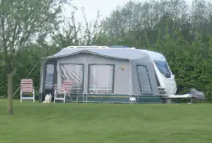 Wohnwagen mit Vorzelt auf einem Campingplatz, zwei Campingstühle und Rasenfläche vor dem Fahrzeug, umgeben von Bäumen.