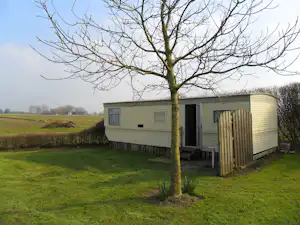 Mobilheim auf grünem Rasen mit Holzveranda, seitlichem Eingang und Baum im Vordergrund, im Hintergrund Felder und Hecken.