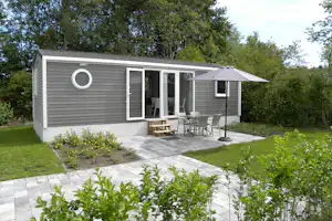 Mobilheim mit Terrasse, Marktschirm und Gartenmöbeln vor gepflastertem Weg, umgeben von Bäumen und Grünpflanzen.