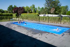 Campingpool mit blauer Wasserfläche, Einstiegshilfe und Personen im Becken, im Hintergrund Wohnwagen und Zäune im Grünen.