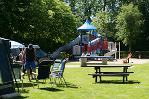 Spielplatz mit Rutsche und Klettergerüst auf dem Campingplatz, daneben Wohnwagen, Auto und Campingstühle im Grünen.