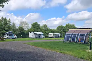 Wohnmobile und ein Campingzelt auf einem gepflegten Stellplatz, mit Grasfläche, Bäumen und Schotterweg im Hintergrund.