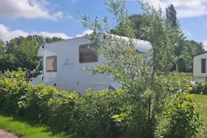 Wohnwagen mit Fenster und Markenzeichen, davor Olivenbaum und Hecken, im Hintergrund weitere Mobilheime und Bäume.