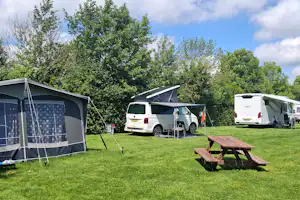 Wohnmobil-Stellplatz mit Vorzelt, Picknicktisch und mehreren Fahrzeugen auf grünem Rasen, umgeben von Bäumen.