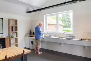 Gemeinschaftsküche im Campinghaus mit Spülbecken, Wasserhahn, Waschmaschine und Fensterfront, daneben ein Holz-Tisch.