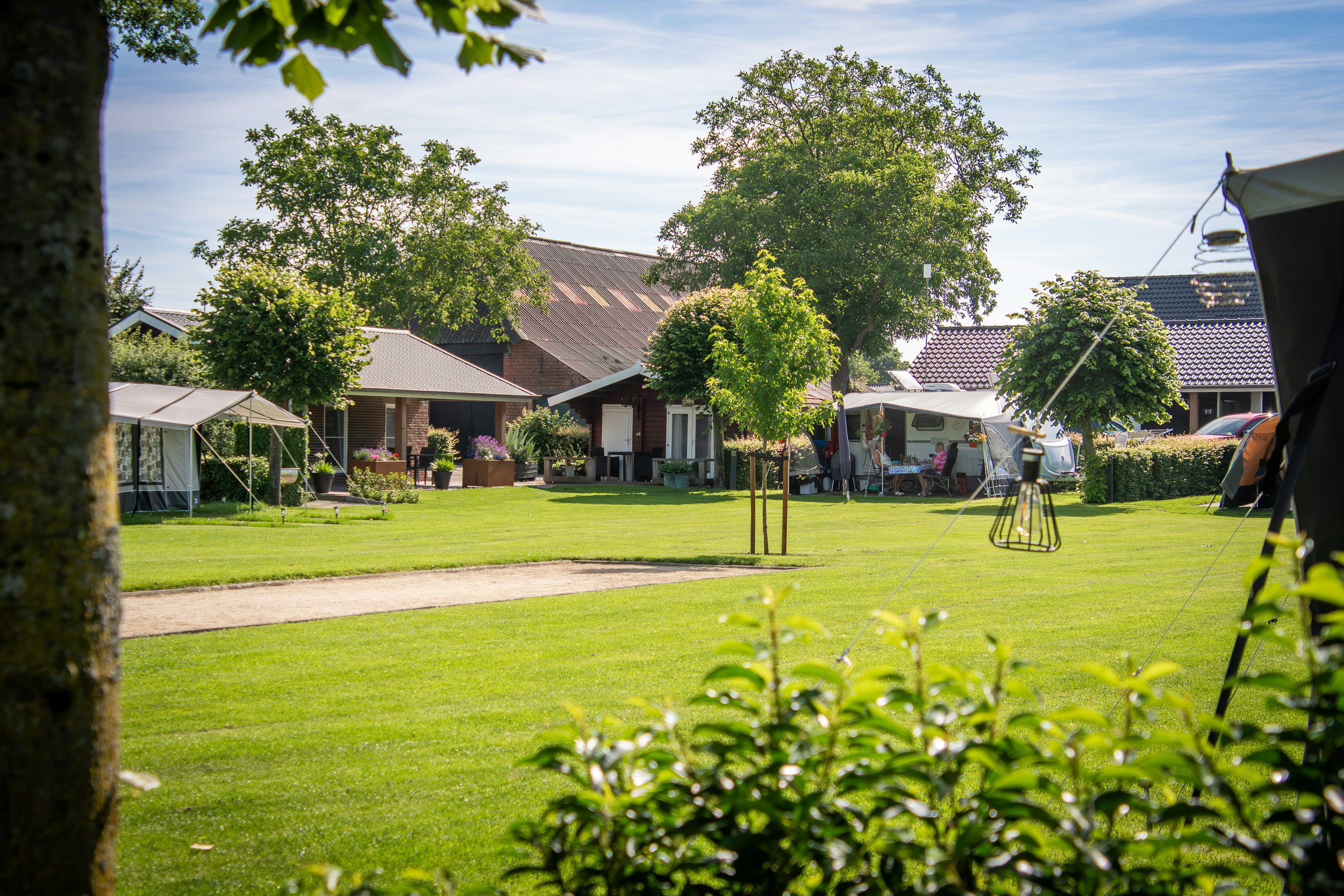 Gepflegter Campingplatz mit Rasen, Bäumen und Zelten, im Hintergrund Gebäude und ein Wohnwagen unter Markisen.