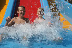 Zwei Kinder rutschen auf einer blauen und roten Wasserrutsche, Wasser spritzt, sie schauen nach vorn.