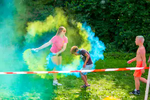 Kinder laufen im grünen Park und sprühen farbiges Pulver aus einer roten Absperrung, umgeben von Bäumen.