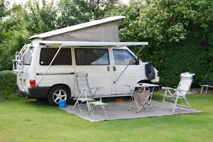 Weißes Wohnmobil mit Markise, davor Campingstühle und Tisch auf grünem Rasen, umgeben von Hecken und Bäumen.